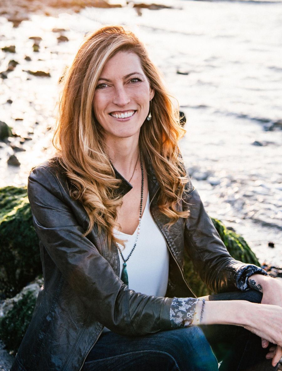 Alana Joy Newton sitting on driftwood at the shoreline, smiling toward the camera